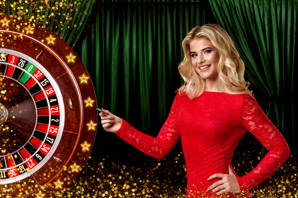 A smiling blonde woman in a lacy red dress stands next to a roulette wheel on a stage. Behind her is a green velvet curtain. Where can I play live dealer games in Michigan? Is it safe to play these games at MI online casinos? Are they fair? 