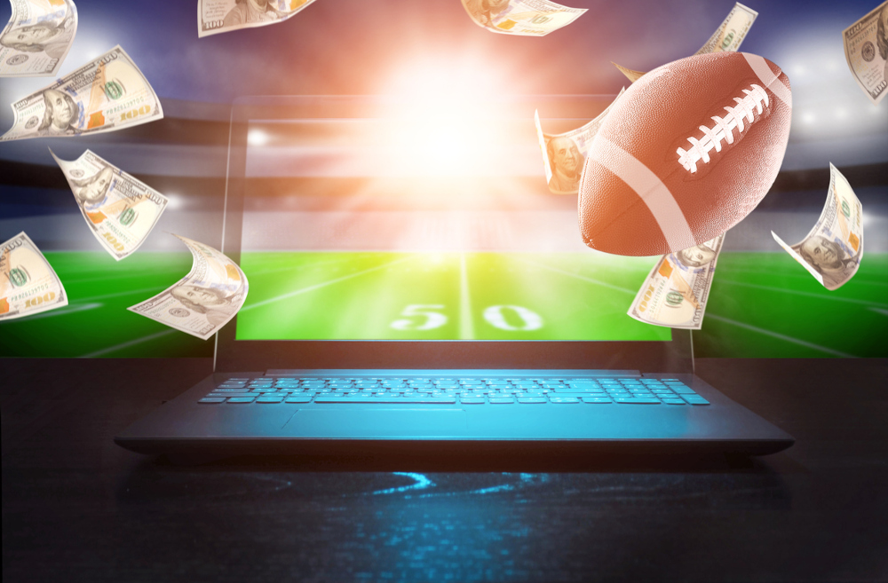 a 3d rendering of a laptop with a football field on the screen and behind it. a football is flying towards the computer as money flies around. representative of online sports betting.