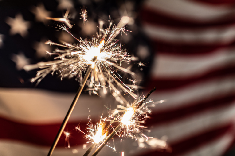 an american flag is seen on the left. next to it, two hands holding sparklers reaching towards each other in the middle of the frame in front of a dark nighttime background.